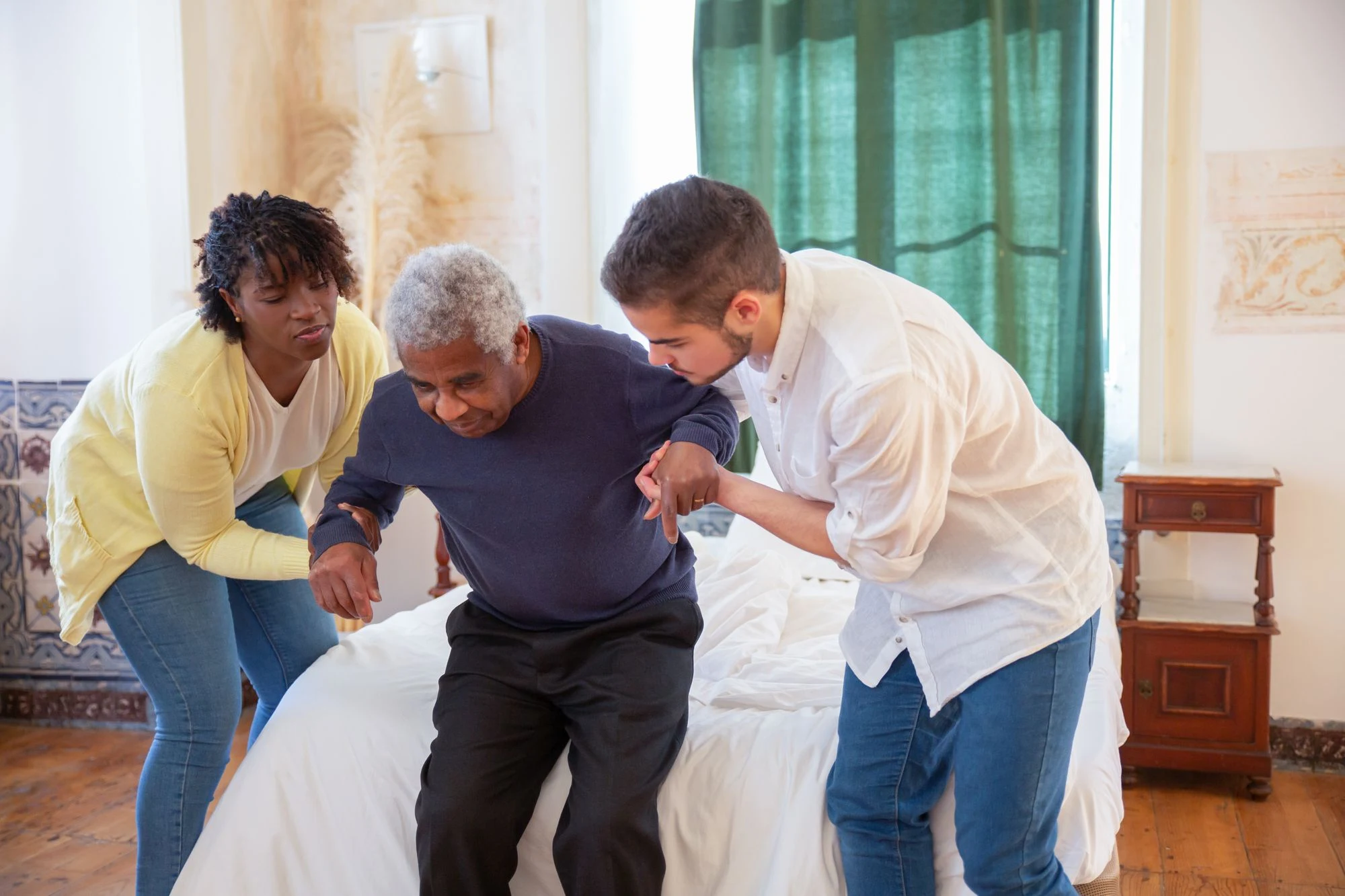 Two people assisting an older adult to stand at home.
